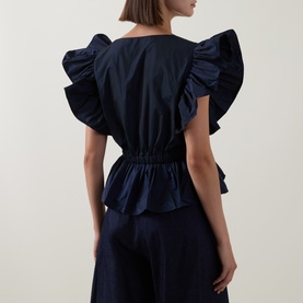 Model wearing Roisin Linnane Navy Top, back view, with dramatic ruffle sleeves, elasticated waist, and styled with navy wide-leg trousers.