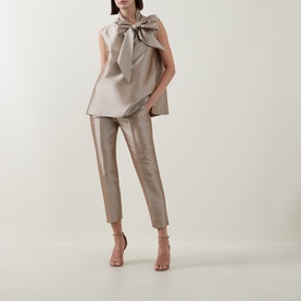 Model wearing Roisin Linnane Twig Cropped Slim-Leg Trousers in gold satin finish, styled with a matching bow-detail sleeveless top and strappy heels.