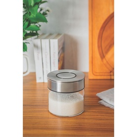 Tramontina clear glass storage container with stainless steel lid, styled on a wooden surface with books and plant in background.