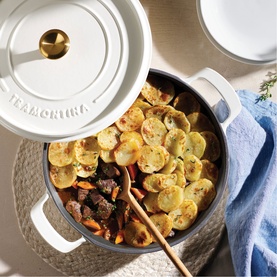 Tramontina latte cast iron pan with lid, filled with sliced potato and vegetable stew, styled on a rattan mat with blue linen