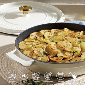 Tramontina latte ceramic pan filled with sliced potato bake, with gold knob lid resting alongside, on neutral surface