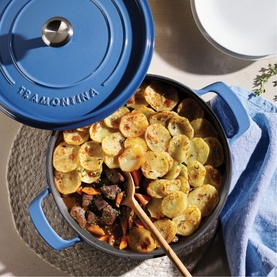 Tramontina blue cast iron casserole pan with lid, filled with roasted potato and vegetable bake, styled on a woven mat