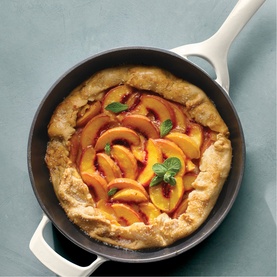 Tramontina latte skillet with rustic peach galette, golden pastry and fresh mint garnish, on a teal surface