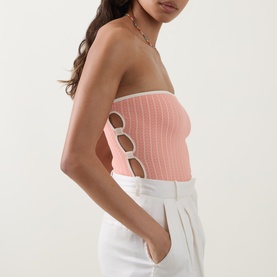 Model wearing Eres pink strapless bandeau swimsuit with cut-out side details and white contrast trim, styled with white wide-leg trousers