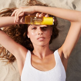 Model with Marc Jacobs Daisy Mist Lemon Spritz Hair and Body Mist, holding yellow bottle, lying on sand in white swimwear