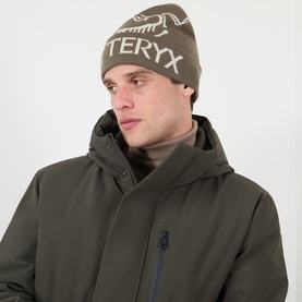 Model with Arcteryx Bird Word Toque Beanie Hat in Mongoose/Sea Salt, worn with olive hooded jacket. Knit hat features contrast logo lettering.