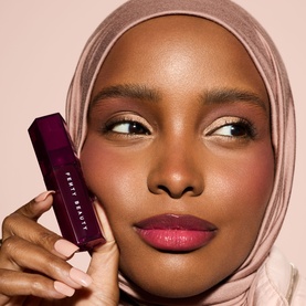 Model wearing Fenty Beauty Gloss Bomb Burst Colour-Intensifying Lip Luminiser in Pink Blink, holding deep plum tube, neutral glam makeup look