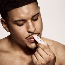 Model with Fenty Skin lip care product, applying balm to lips, shirtless against neutral background
