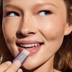Model with Fenty Skin lip care product applied to lips, holding lilac-coloured stick applicator close to mouth