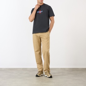 Model wearing Gramicci Ladybug Logo T-Shirt in black with chest graphic, styled with beige chinos and grey trainers.