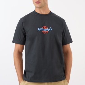 Model wearing Gramicci Ladybug Logo T-Shirt in black with chest graphic, crew neck, short sleeves, paired with beige trousers.