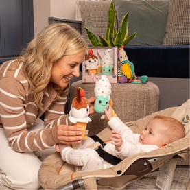 Play Easy multi-colour plush toy held by smiling woman entertaining baby in bouncer chair, with additional soft toys visible in background