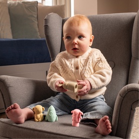 Baby seated in grey armchair holding a small toy, with colourful pastel soft toys arranged nearby on the cushion