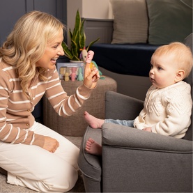 Woman showing a small pastel rainbow soft toy rattle to a baby seated on a grey sofa in a cosy living room setting