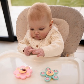 Baby in high chair exploring two silicone spinner toys in pink and mint green flower shapes on white tray