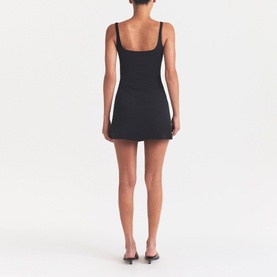 Model wearing Skims black mini dress, rear view, with thin shoulder straps, styled with brown heeled mules.