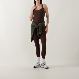 Model wearing dark brown bodysuit and matching leggings, styled with olive shirt tied at waist, white socks and multicolour trainers