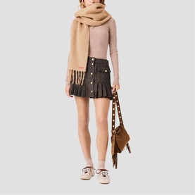 Model wearing Maje mini skirt with pleated hem and button detail, styled with camel scarf and ribbed top, carrying suede fringed bag