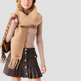 Model wearing Maje scarf in camel with fringe detail, styled with beige roll-neck, dark mini skirt, and tan fringed shoulder bag.