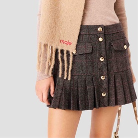 Model wearing Maje mini skirt in dark multi tweed with pleated hem and gold buttons, styled with beige fluffy scarf