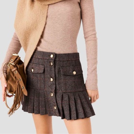 Model wearing Maje multi tweed pleated mini skirt with gold buttons, styled with beige knit top, camel jacket and tan fringed bag