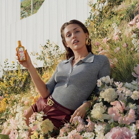 Model holding Gucci EDP bottle, seated among blooming flowers, wearing grey polo and burgundy trousers with gold hoop earrings.