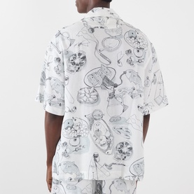 Model wearing Jacquemus Fish Graphic Bowling Short in off-white with all-over monochrome food and fish print, notched collar, short sleeves.