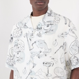 Model wearing Jacquemus Fish Graphic Bowling Shirt in off-white with blue fish print, notched collar, short sleeves, styled with gold chain necklace.