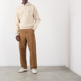 Model wearing Jacquemus beige cotton hoodie with small logo, styled with tan wide-leg trousers and white trainers.