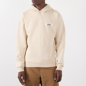 Model wearing Jacquemus beige cotton hoodie with small logo patch on chest, styled with tan trousers.