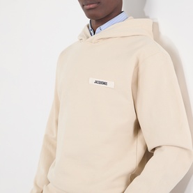 Model wearing Jacquemus beige cotton hoodie with small logo patch on chest, styled with light blue shirt underneath.