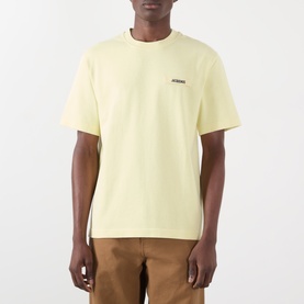 Model wearing Jacquemus The Gros Grain Logo T-Shirt in pale yellow, round neck, chest logo patch, styled with tan trousers.