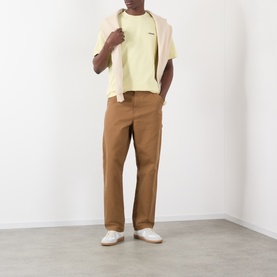 Model wearing Jacquemus The Gros Grain Logo T-Shirt in pale yellow, styled with tan trousers and a cream overshirt.