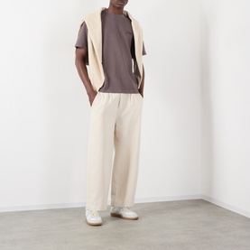 Model wearing Jacquemus brown cotton T-shirt with cream wide-leg trousers, cream gilet draped over shoulders, and white trainers.