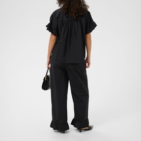 Model wearing Kaffe Black Deep ruffle-hem wide-leg trousers, styled with matching ruffle blouse, small black bag and heels, shot from behind.