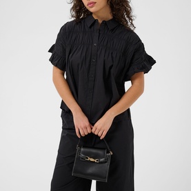 Model wearing Kaffe black deep short sleeve shirt dress with ruched bodice and frill sleeves, styled with black trousers and mini bag