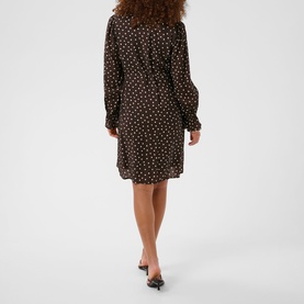 Model wearing a black mini shirt dress with white polka dot print, long sleeves and belted waist, styled with black heeled mules.