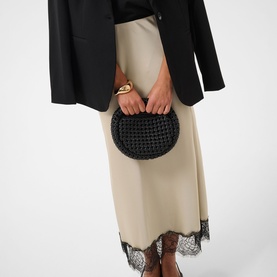 Model carrying a black woven half-moon clutch bag, styled with a black blazer and beige lace-hem midi skirt