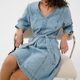 Model wearing light blue denim V-neck mini dress with tie waist belt and rolled sleeves, seated on cream chair