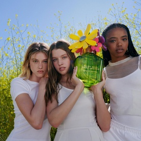 Three models in white outfits in a sunny meadow, one holding Marc Jacobs Daisy Wild Eau So Extra Eau de Parfum green floral bottle