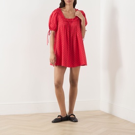 Model wearing Damson Madder Jemima Cherry Mini Dress in red cherries, featuring scoop neckline, puff sleeves with tie cuffs, styled with black flat