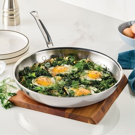 Hestan ProBond Luxe Skillet 28cm in polished stainless steel, shown on wooden board with fried eggs and greens, marble background.