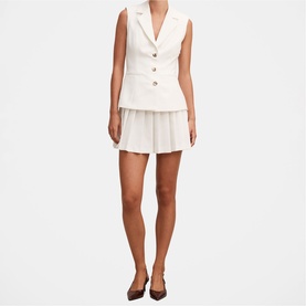 Model wearing white sleeveless blazer vest with gold buttons and matching pleated mini skirt, styled with black heels
