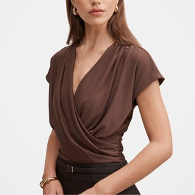 Model wearing brown short-sleeve wrap-front top with draped V-neckline, styled with a dark belted skirt and gold jewellery