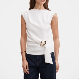 Model wearing white sleeveless top with asymmetric tie detail and gold-tone ring hardware, styled with dark blue jeans and a gold bracelet.