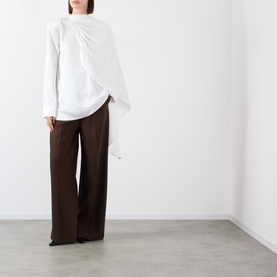 Model wearing white asymmetric draped long-sleeve top with flowing side panel, styled with wide-leg dark brown trousers