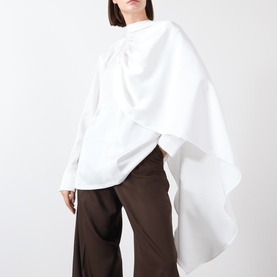 Model wearing white oversized draped cape top with asymmetric hem, styled with wide-leg brown trousers