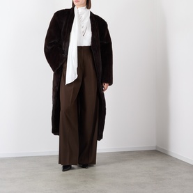 Model wearing a dark brown faux fur longline coat, styled with a white tie-neck blouse and wide-leg brown trousers