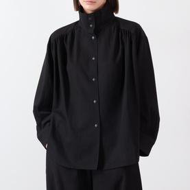 Model wearing black gathered high-neck long-sleeve shirt with button-front placket, styled with wide-leg black trousers