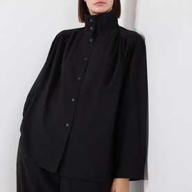 Model wearing black high-neck button-through oversized shirt with draped shoulders, styled with matching black wide-leg trousers